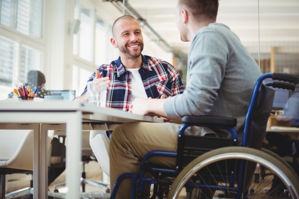 This is an image of a man in a wheelchair at a desk. he is talking to another man. They are in a meeting. Both men are in smart casual dress and are in, what looks like their 30s. Both have short hair. They are in a room which looks like a modern office.