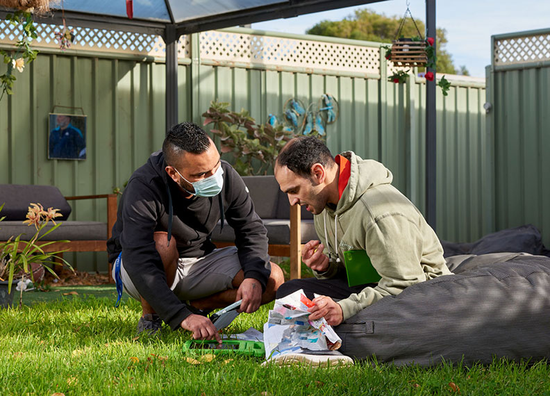 A disability support worker is crouching down to talk to a man who is sitting outdoors on a beanbag