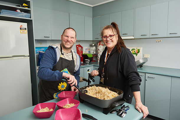 A man and a woman stand at a kitchen bench, the man is stirring food in a large electric frying pan, they are both smiling