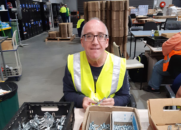 A man is wearing a high-vis vest and sitting at an assembly workstation in a warehouse