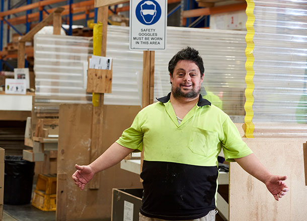 A man in high vis work wear is standing in a warehouse, he is holding his arms out and smiling