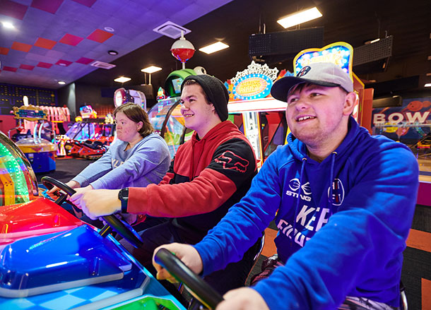 Three people sit side by side on a car racing game in an arcade