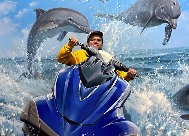 A man poses in a set, it appears he is riding a jet ski with dolphins leaping over him