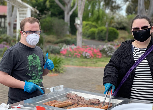 Two people are standing at a BBQ in the park, they are wearing facemasks, gloves and holding tongs to cook meat on the grill