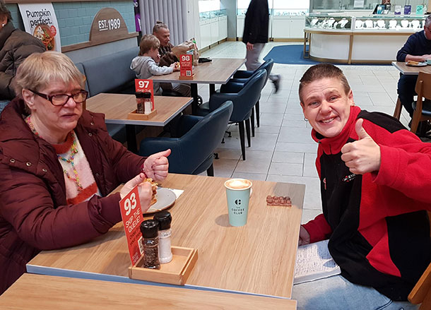 Two people are sitting at a cafe table eating lunch, one has their thumb up and is smiling