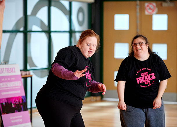 Two women wearing t-shirts that say BeatFreakz in a dance studio, one is dancing the other smiles standing beside her