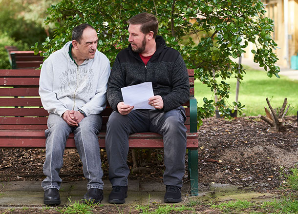 Two men sit outside on a bench talking to each other