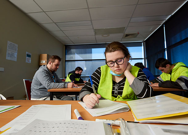 Caitlin is sitting at a desk in a classroom, she is looking down at her classwork and writing something