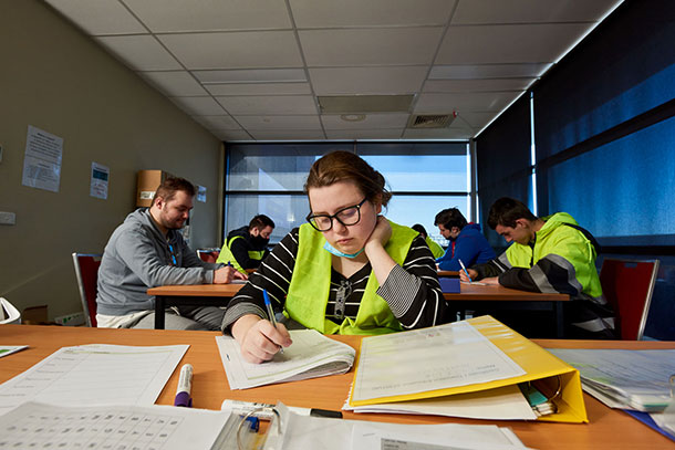 Caitlin is sitting in a classroom, she is holding a pen and is looking down reading her worksheet, there are folders and papers on the desk, behind her other SLES participants are working at desks