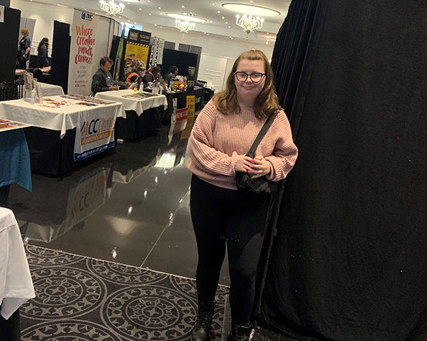 Caitlin stands in front of a row of expo stalls, she is smiling