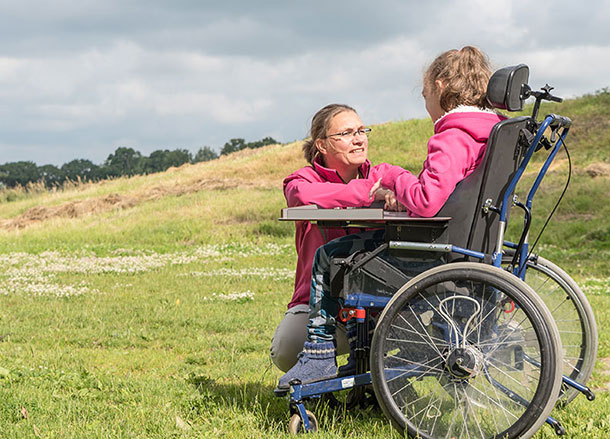 A carer squats down to speak to someone in a wheelchair, they are outside on a grassy hill