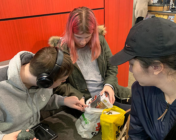 Three people are sitting on some benches, they are looking into shopping bags, one person is pulling a POP collectible out of a bag