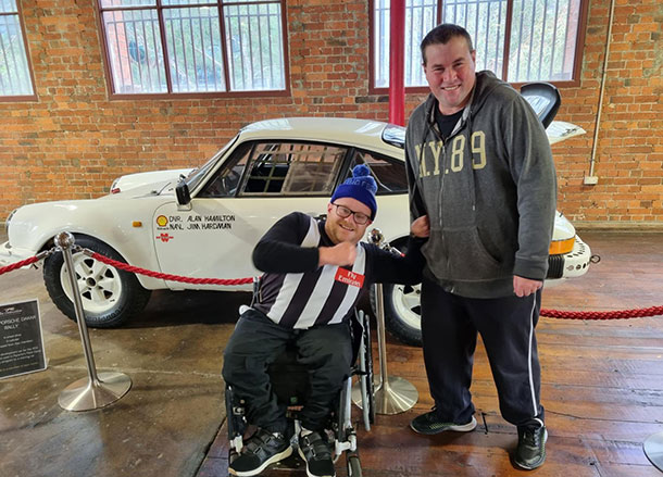 Two men one in a wheelchair stand in front of a classic car on display