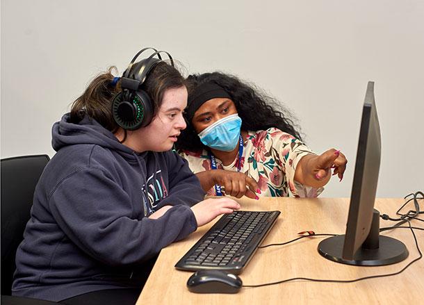 A young woman is wearing headphones and sitting in front of a desktop computer, a facilitator is beside her pointing to something on the monito