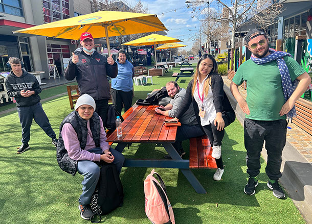 Seven people sit and stand around an outdoor dining table in a sunny street mall