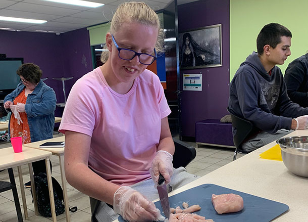 A woman is standing at a bench chopping raw chicken on a chopping board