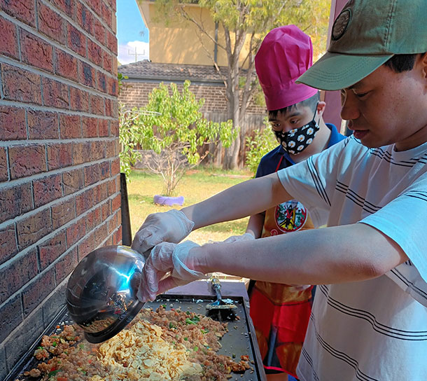 Two men stand in front of an outdoor grill, one is wearing a red chef's hat, the other is pouring ingredients from a bowl onto the grill