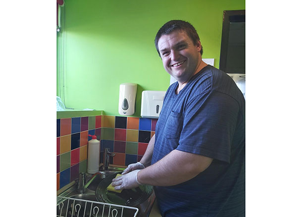 Daniel stands at the sink washing dishes, he is smiling