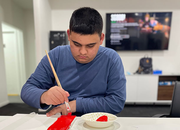 A man is sitting at a table holding a paintbrush and painting a red stripe on a canvas