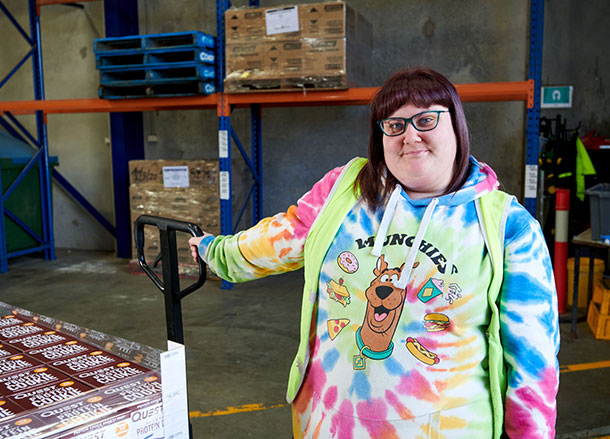 A woman in a tie-die shirt with a high vis vest over it stands standing with a hand on a pallet mover in a warehouse