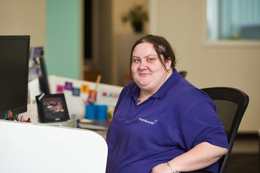 Dee-Jay is wearing a purple Mambourin-branded polo shirt, she is sitting at the reception desk smiling