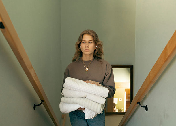 A young woman is wearing earbuds, while she carries a pile of folded towels and walks up stairs