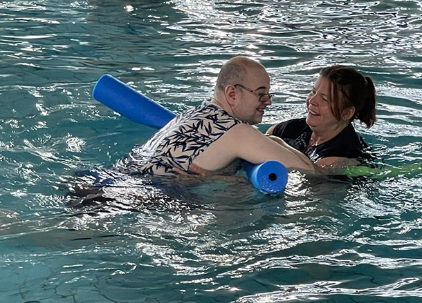 A man is in a pool leaning on a pool noodle with a support worker assisting 