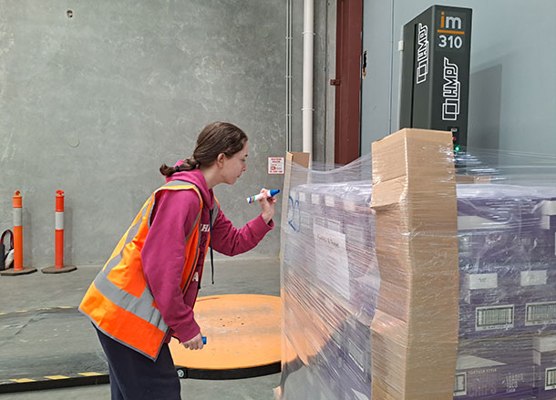 A woman in a high-vis vest is using a marker to label a pallet in a warehouse