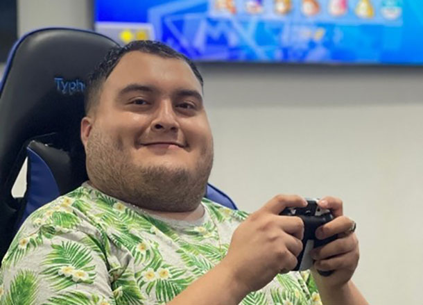 A man is smiling, he is sitting in a gaming chair and holding a video game controller in his hands, a large screen is behind him