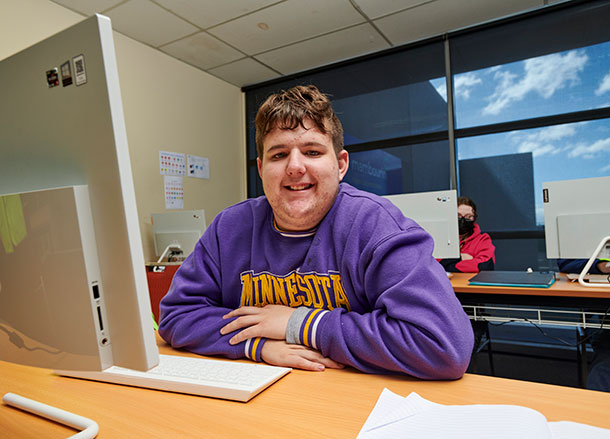 Ethan is sitting in front of a computer in a classroom, he is smiling