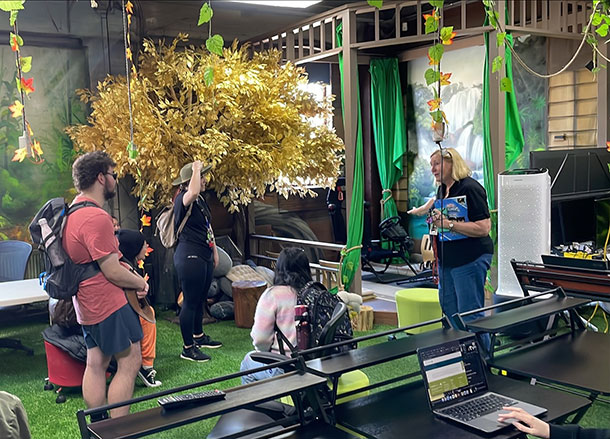 A group of people stand listening to a woman talk, they are in a room with a small stage, work stations, fake grass and fake trees