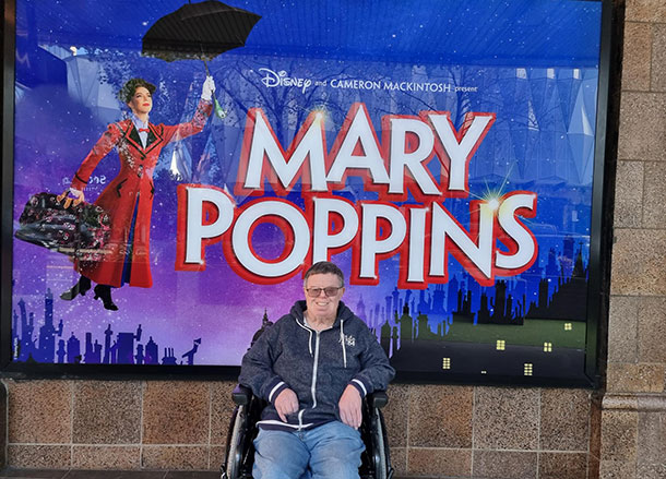 A man is seated in a wheelchair outside a theatre poster for Mary Poppins