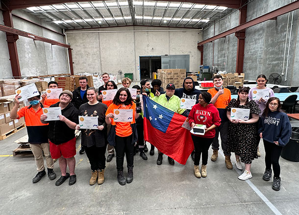 A group of people stand together in a warehouse, they are all holding certificates and awards