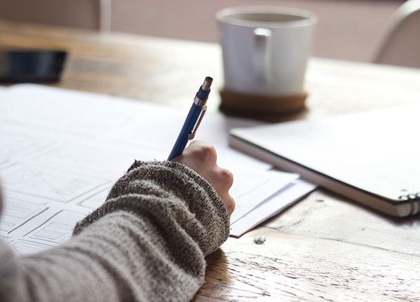 An arm resting on a table, writing notes on a piece of paper on a table, a notebook and a coffee mug are also on the table