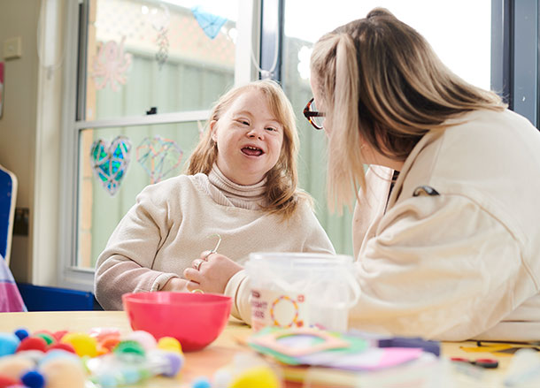 A woman is looking at her support worker smiling, there are craft supplies on the table between them