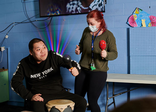 A man is sitting playing a drum, a support worker stands beside him shaking a maraca