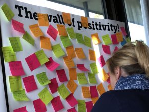 This is a picture of a white wall covered in post-its. The white wall has the words Wall of Positivity in heavy black writing. The post-its each have a positive message on them about what it's like working at Mambourin. In the image you can see the back of a woman's head as she is placing her post-it on the wall. She has blonde hair in a ponytail.