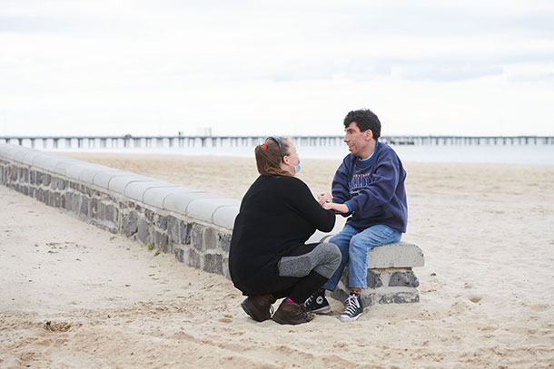 A support worker crouches down to talk to the person she is supporting who is sitting on a low wall at the beach