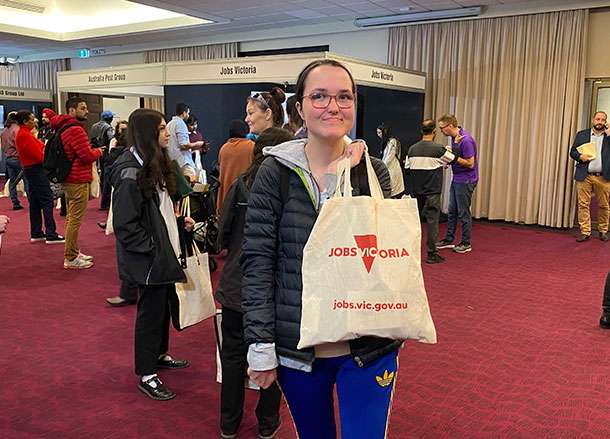 A young woman holds up a Jobs Victoria tote bag in front of a booth