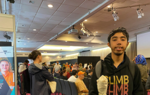 A person stands smiling, holding a tote bag under his arm, behind him people are seated as though watching a presentation