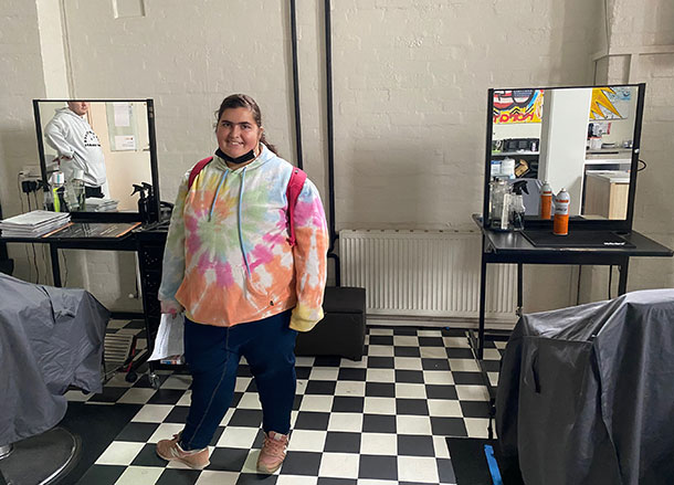 A young woman stands in a room set up for a Barber training course, there are hairdressing stations with mirrors and a black and white tiled floor