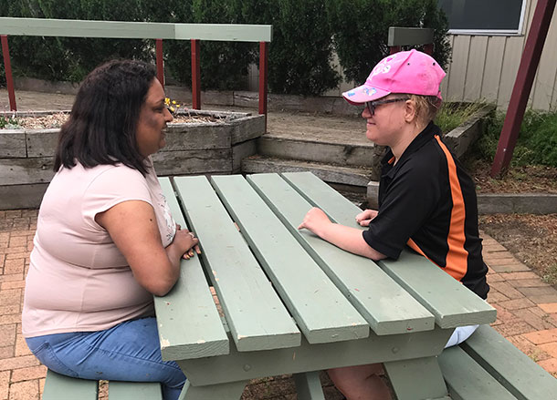 Two people sit either side of an outdoor table, they are smiling and looking at each other