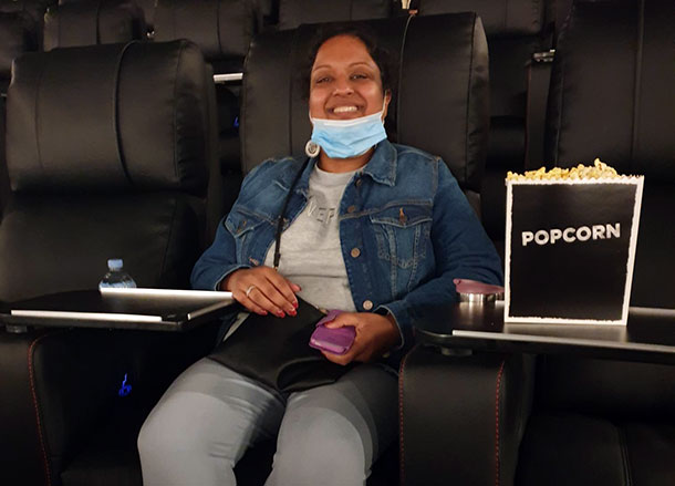 A woman sits in a cinema, she has a box of popcorn beside her and she is smiling