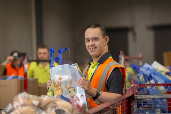 This is an image of a man working in a warehouse. He is wearing a high vis vet in orange. He is in his mid 20s and has black short hair. he is holding out a showbag he has just filled with merchandise and he is smiling at the camera.