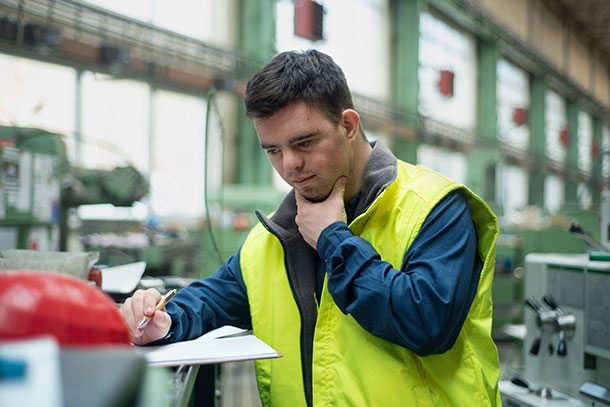 Young man with Down Syndrome filling in application form in a warehouse, he he is raising his hand to his chin as if thinking or stressed