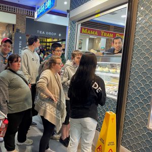 Seven people stand near a food counter talking to the server