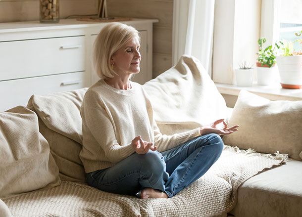 A woman is sitting cross-legged on a couch with her hands resting on each knee and her eyes closed