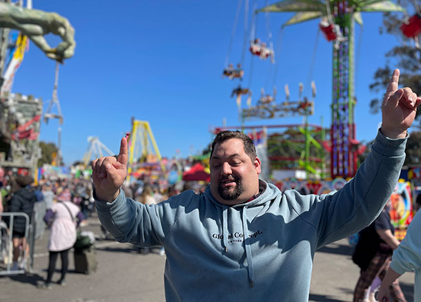 A man stands holding up his arms in happiness, showround attractions are visible behind him, it is a sunny day with a bright blue sky