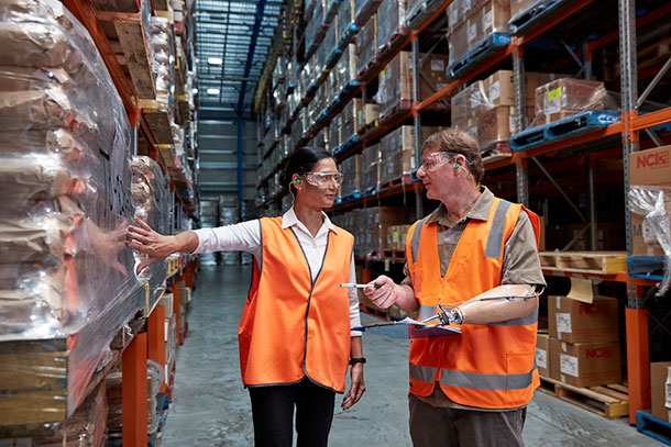 Two people in high-vis vests stands in a warehouse talking, one of them has a prosthetic arm