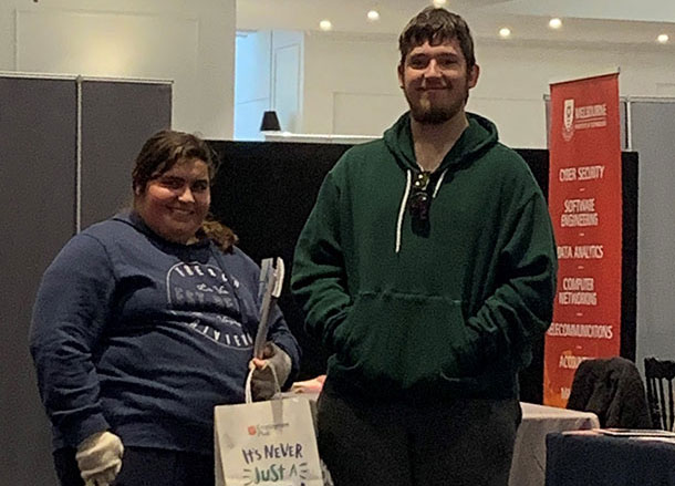 A young woman and a young man stand side by side, they are in an event space, a stall with a banner is behind them